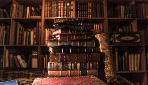 Stack of antique books on a table, with shelves full of volumes in the background of a warm library.