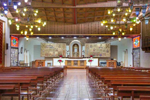  Interior de una iglesia con bancas de madera alineadas, lámparas colgantes y un altar decorado con imágenes religiosas y flores al fondo.
