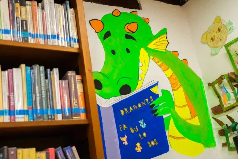 Mural colorido de un dragón verde leyendo un libro en una biblioteca, junto a estanterías llenas de libros que aportan un ambiente creativo y educativo.
