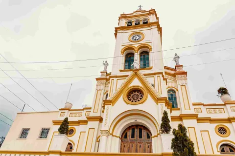 Primer plano de la fachada de una iglesia color marrón claro con toques de color amarillo. Hay algunos pinos pequeños a los lados.