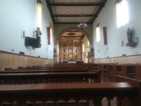Interior de una iglesia con bancas de madera, paredes blancas y un altar dorado al fondo, iluminado por luz natural que entra por ventanas laterales.