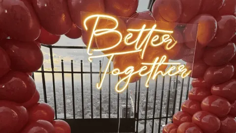  Estructura decorativa con globos rojos enmarca un letrero luminoso que dice “Better Together”. Al fondo, se aprecia una vista urbana desde gran altura tras una baranda.