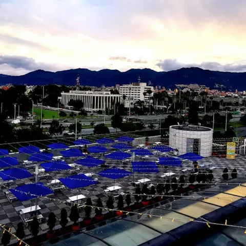 Panoramic view of Bogotá at sunset, with mountains in the background and an urban terrace with blue umbrellas in the foreground.