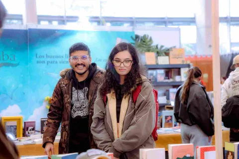 Two visitors pose and explore a book display booth at a literary fair, in the middle of a lively cultural atmosphere packed with readers, publishers, and reading-centered activities.