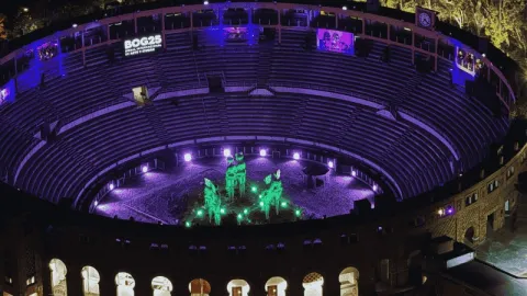 Vista nocturna de una plaza de cultural iluminada con luces moradas y verdes, donde una instalación artística ocupa el centro. Las gradas circulares rodean el espacio, creando un ambiente escénico y vibrante.