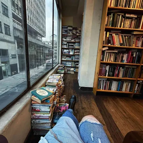 View from a bookstore with shelves and stacks of books next to a window, while a person rests looking out at the street.