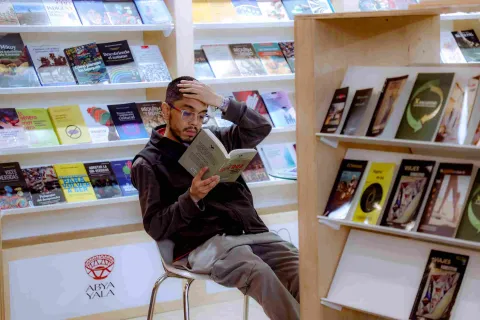A visitor rests and reads a book intently in front of shelves filled with new releases and editorial publications at a literary fair.