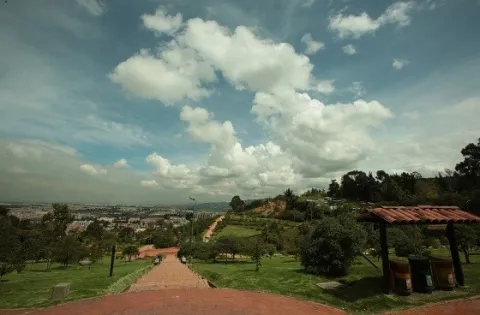 From the top of Mirador de los Nevados Park, a vast green landscape unfolds, sloping down towards the city, framed by natural trails and a sky filled with clouds that highlights the tranquility of the place.