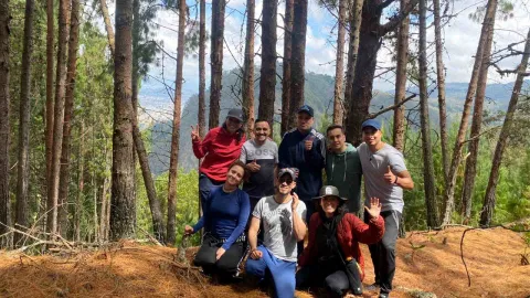 Grupo de personas posa sonriente en un bosque de pinos, rodeado de montañas y vegetación, durante una salida al aire libre.