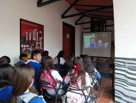 Grupo de estudiantes sentados en sillas dentro de un museo, observando una proyección en pantalla de una animación. La sala tiene paredes blancas, vigas de madera en el techo y un afiche ilustrado colgado en la pared lateral.