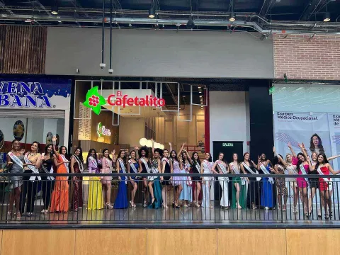 A group of pageant contestants pose inside a shopping mall, dressed in elegant suits and wearing sashes.