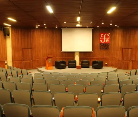  Auditorio Rogelio Salmona del Centro Cultural Gabriel García Márquez, con filas de sillas para el público frente a un escenario equipado con pantalla de proyección, atril y cuatro sillones. El recinto destaca por sus acabados en madera y su disposición para conferencias, conversatorios y actividades culturales.