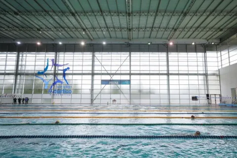  Vista interior del Complejo Acuático de Bogotá, con una amplia piscina de competición de múltiples carriles donde nadadores entrenan bajo una moderna estructura cubierta e iluminada con luz natural.