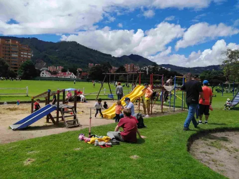 Niños y familias disfrutan de una zona de juegos infantiles y amplias áreas verdes en un parque, con los cerros orientales y un cielo parcialmente nublado al fondo.