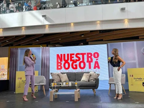 Two women participate in a talk on stage inside the Nuestro Bogotá shopping mall, in front of a screen and an audience.