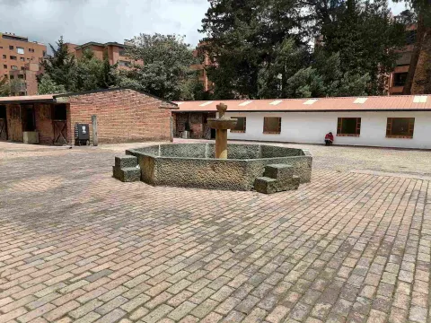 Patio adoquinado con una fuente de piedra en el centro, rodeado por edificaciones de estilo tradicional, árboles altos y edificios residenciales al fondo.