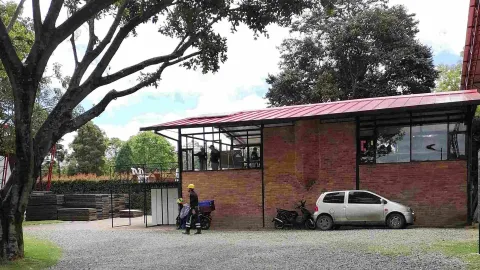 Edificación de ladrillo con cubierta roja en una zona verde, junto a un área de parqueadero y rodeada de árboles de gran tamaño.