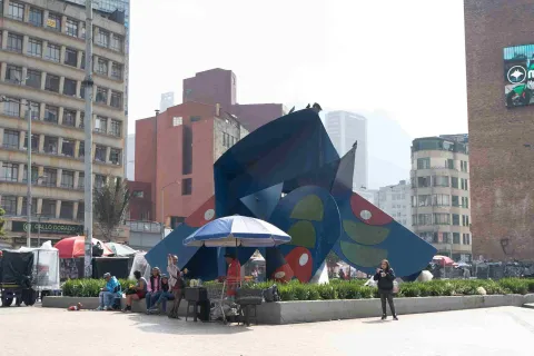 Escultura urbana de gran formato en una plaza del sector de San Victorino, rodeada de zonas verdes, vendedores y peatones. Al fondo se observan edificios del centro de Bogotá, reflejando el ambiente comercial, cultural y cotidiano de esta emblemática zona de la ciudad.