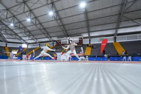 Athletes compete in a fencing match inside the Palacio de los Deportes in Bogotá, on a piste surrounded by stands and competition areas.