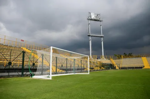 Arco de fútbol sobre la cancha del Estadio de Techo, con graderías amarillas, torre de iluminación y un cielo oscuro y nublado al fondo.