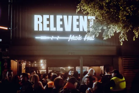  Fachada iluminada de Relevent Music Hall durante la noche, con un grupo de personas reunidas en la entrada bajo el letrero luminoso del establecimiento.