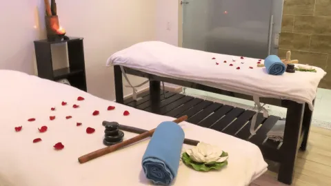 Sala de masajes con dos camillas decoradas con pétalos de rosa, toallas, piedras y elementos de relajación en un ambiente cálido y tranquilo.