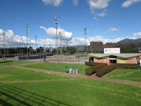 View of the tennis courts at the High-Performance Center in Bogotá, surrounded by green areas, sports lighting, and city buildings in the background.