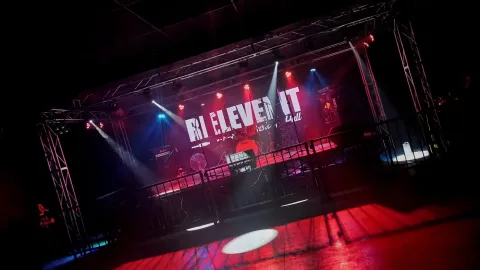  Escenario de Relevent Music Hall con batería, micrófonos e iluminación roja, azul y blanca, listo para la presentación de una banda en vivo.
