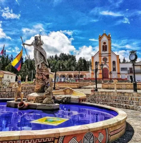 Fuente con esculturas y un monumento central en una plaza, frente a una iglesia de fachada blanca y amarilla, bajo un cielo azul con nubes.
