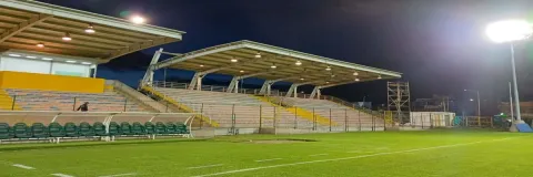  Vista del Estadio Olaya Herrera durante la noche, con la cancha iluminada, las graderías cubiertas y el banco de suplentes en primer plano.