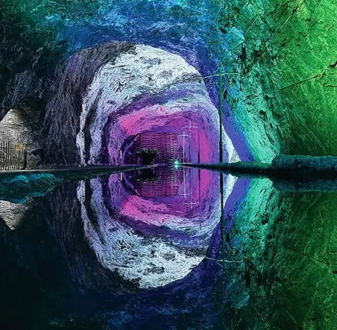 Interior de una mina de sal iluminada con luces verdes, azules y moradas, cuyo reflejo en el agua crea un efecto visual simétrico.