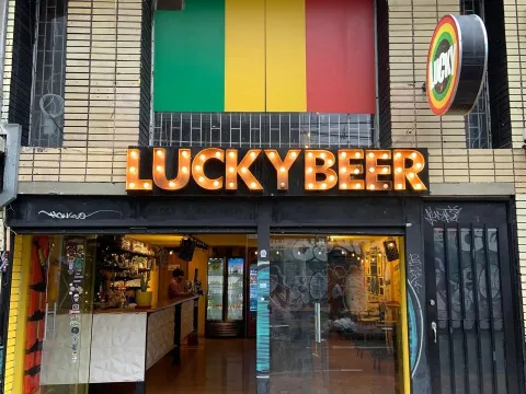 Fachada de Lucky Beer con un letrero luminoso, una bandera con colores verde, amarillo y rojo sobre la entrada, y un bar visible a través de sus puertas de vidrio.