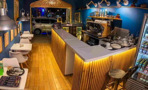  Interior de una cafetería con barra de madera iluminada, máquina de café, mesas blancas y decoración en tonos azules, vista desde la entrada durante la noche.