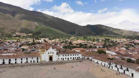 Vista panorámica de la Plaza Mayor de Villa de Leyva, rodeada de casas coloniales de techos de teja, con la iglesia principal y montañas verdes al fondo. Interior de una iglesia colonial con bancas de madera, un arco decorado con detalles dorados y rojos, y un altar principal ornamentado al fondo.