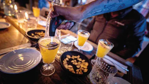 Mano usando un soplete pequeño para flamear una rodaja de fruta sobre un cóctel amarillo en vaso de cristal.