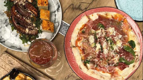 Vista superior de una mesa de madera con una pizza de jamón, un plato de pulpo, un cóctel y aperitivos variados.
