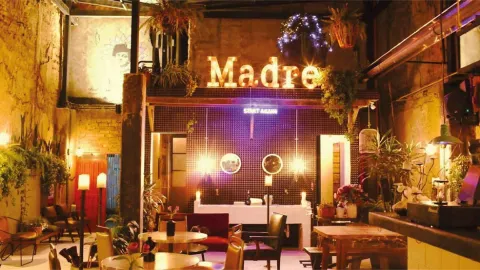 Interior de estilo rústico e industrial con luces cálidas, plantas y un letrero luminoso con la palabra Madre.