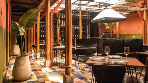 Comedor del restaurante con piso de baldosas coloridas, columnas de madera, cava de vinos y cocina abierta al fondo.