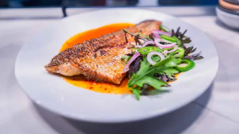 Filete de pescado dorado a la parrilla servido con ensalada de cebolla morada y ají sobre un plato blanco.