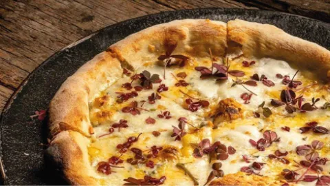 Acercamiento a una pizza artesanal con queso fundido y brotes morados sobre una fuente sobre mesa de madera.