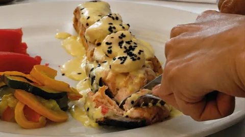 Una persona usa un tenedor y un cuchillo para cortar una pieza de salmón cubierto de salsa y sésamo negro en un plato.