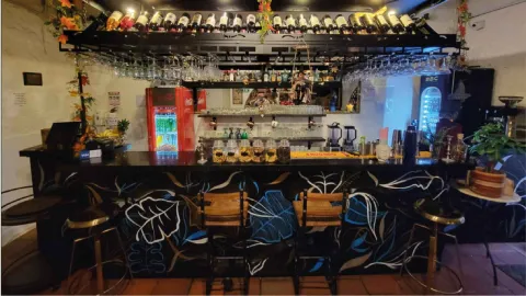 Un bar con una encimera de madera oscura pintada con hojas azules y blancas, y estantes llenos de botellas de alcohol.