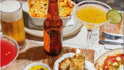 Mesa servida con cócteles, vaso de cerveza, botella de cerveza, pan artesanal, entrada horneada y pizza.