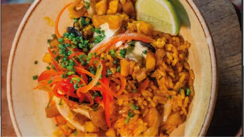 Tazón de arroz preparado con mariscos, cebolla en tiras, verduras picadas y una rodaja de limón al borde.