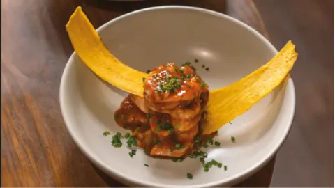 Tazón blanco con mariscos en salsa roja decorados con hierbas y dos crujientes tajadas largas de plátano verde.