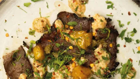 Un primer plano de una pieza de carne asada cubierta con hierbas frescas, copos de chile y una salsa cremosa en un plato.