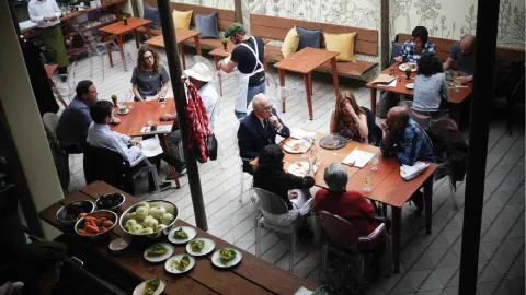 Vista aérea de un patio de restaurante lleno de gente cenando en mesas de madera, con una cocina abierta visible.