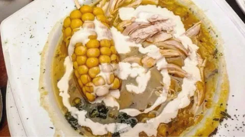Sopa tradicional de ajiaco servida en plato hondo con mazorca, pollo desmechado y crema de leche derramada.