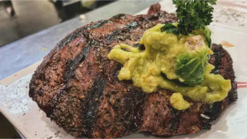 Jugoso corte de carne a la parrilla sobre plato blanco, cubierto con una porción generosa de guacamole y perejil.
