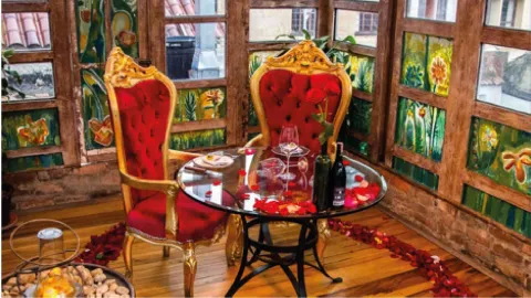 Mesa redonda decorada con pétalos de rosa, vino y dos sillas elegantes de terciopelo rojo junto a un ventanal.